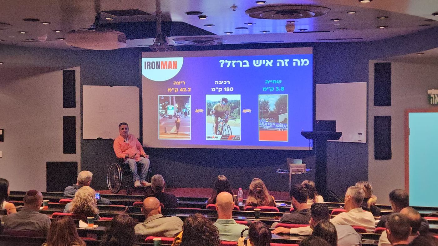 ניר עובדיה מרצה באודיטוריום מלא, על המסך: מה זה איש ברזל? שחייה 3.8, רכיבה 180, ריצה 42.2 ק״מ